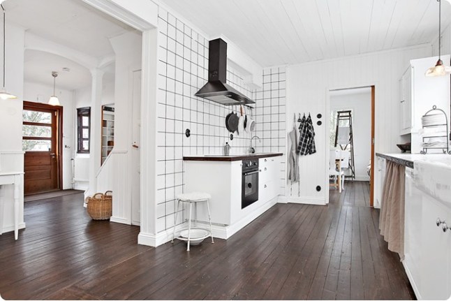 Biała kwadratowa płytka z czarnymi spoinami na ścianie kuchennej z czarnym okapem, brązowa ciemna podłoga z desek na podłodze w białej kuchni