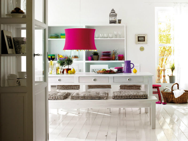 Biały prostokątny stół z szufladami,biała ławka z szarymi siedziskami,białe deski na podłodze i lampa w kolorze fuksji nad stołem