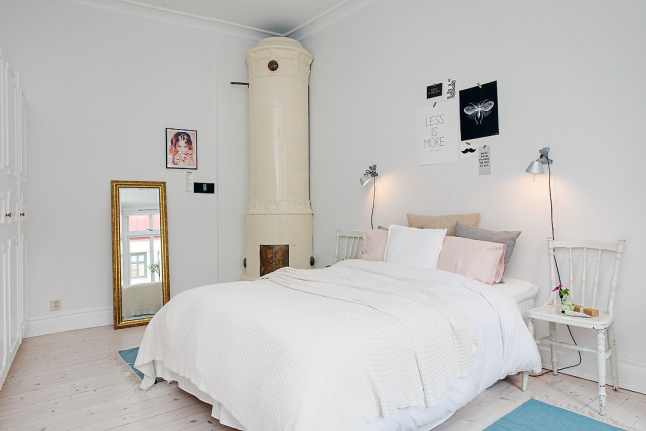 Ceramiczny okragły piec skandynawski,złocone stojace lustro,srebrne lampki przy białym łóżku w sypialni z turkusowymi dywanikami