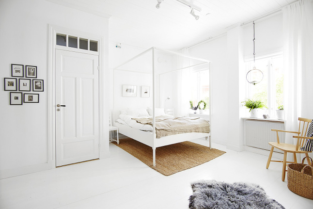 Białe nowoczesne  łóżko z wysokimi ramami i beżową narzutą  , jasnobrązowy dywan i mała galeria fotografii na ścianie