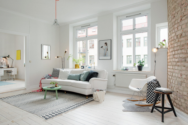 Biała sofa na kółkach,turkusowy stolik pomocniczy kawowy,turkusowe i biało-czarne poduszki,biały fotel na płozach i ściana z czerwonej cegły w salonie,lampa ma czerwonym kablu,