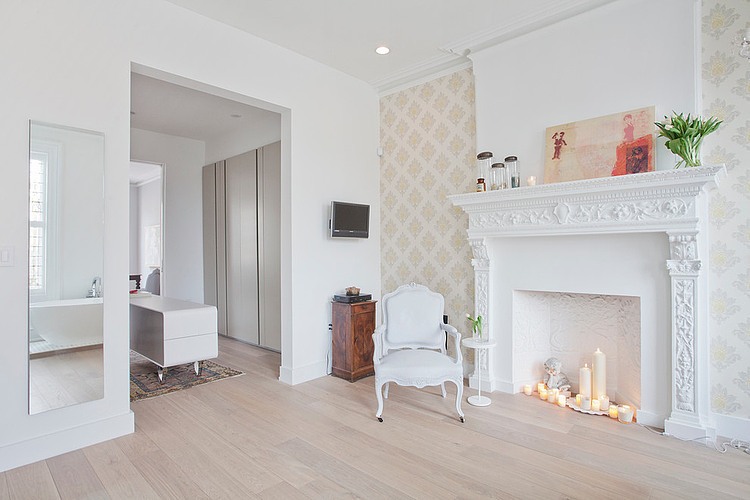Klasyczna tapeta na ścianie,francuskie krzesło i stylowa obudowa kominka ze świecami w aranżacji białej sypialni