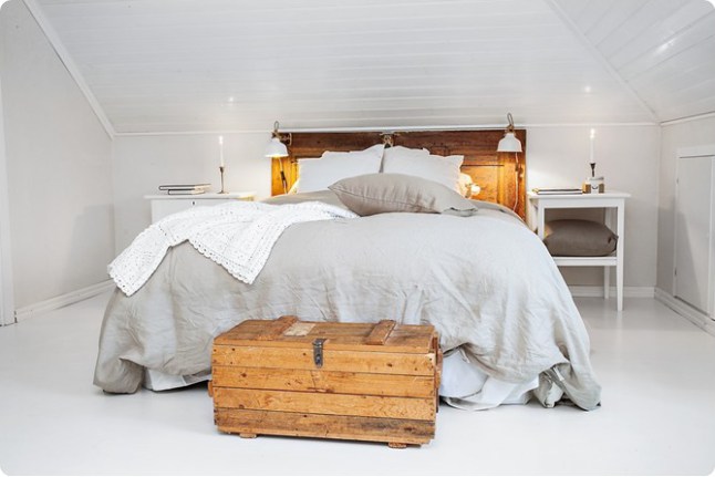 Biała sypialnia na poddaszu z drewnianym łóżkiem, drewnianym miodowym kufrem skrzynią i szarą lnianą pościelą