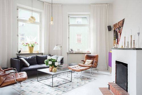 Żarówki na kablu,biały murowany kominek,szara kanapa i nowoczesny skórzany fotel z podnóżkiem w salonie