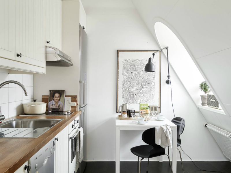 Biała kuchnia z drewnianym blatem, kwadratowy biały stolik z czarnymi krzesłami,grafitowy kinkiet z wysięgnikiem i czarna podłoga w kuchni