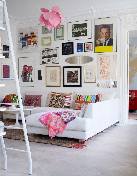 Pomysł na kolorową galerię na białej ścianie z grafik i plakatów i róznobarwne poduszki na sofie w salonie