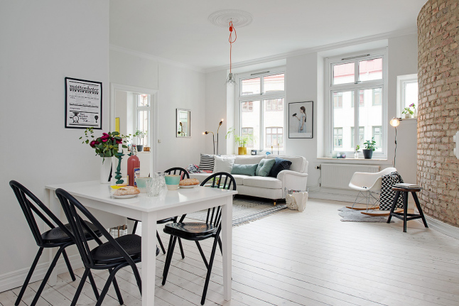 Biały stół z czarnymi krzesłami w otwartej przestrzeni mieszkania z salonem z białą podłogą i ścianą z czerwonej postarzanej cegły