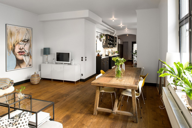 Drewniana podłoga,drewniany stół prostokatny z nowoczesnymi krzesłami,nowoczesny obraz i białe szafki w otwartym salonie z kuchnią i jadalnią