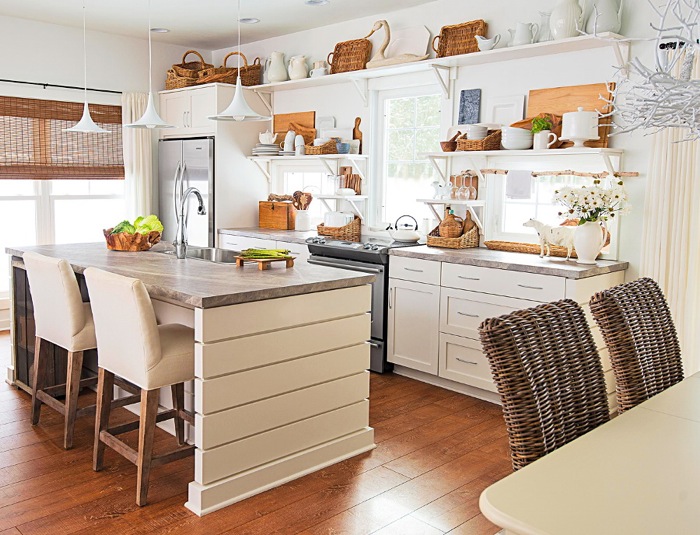 Biała kuchnia z szarymi blatami,kuchenną wyspą i tapicerowanymi wysokimi krzesłami,białe półki wiszące z wiklinowymi i drewnianymi dekoracjami