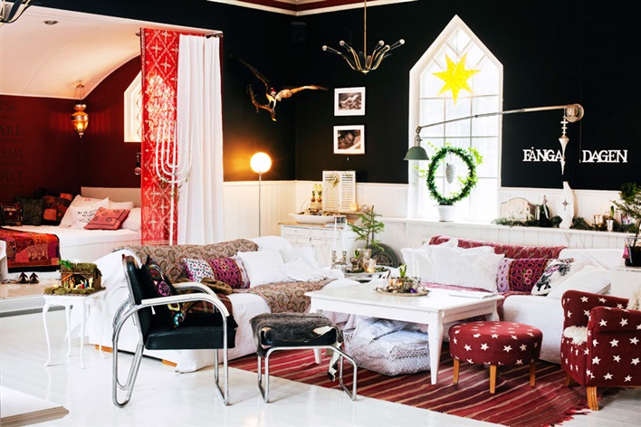 Czerwone i czarne ściany w skandynawskim salonie z biała sofą,czarnymi krzesłami i czerwonym fotelem z podnóżkiem w białe gwiazdki w świątecznej dekoracji