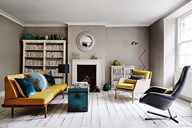 Szare ściany w salonie z kominkiem,żółtą sofą,turkusowym kufrem i białą podłogą z patynowanych desek