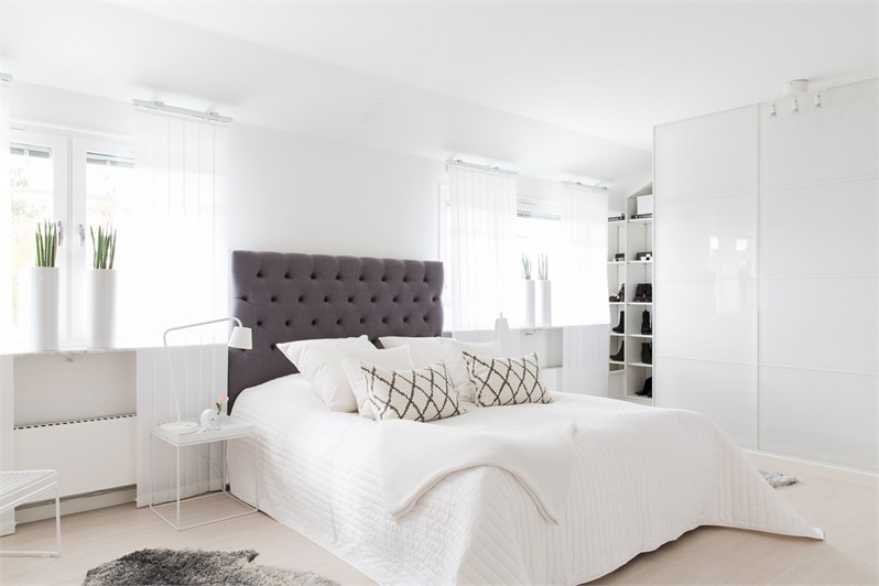Biała sypialnia z czarnym pikowanym zagłówkiem,nowoczesnymi wazonami na oknie i graficznymi klasycznymi poduszkami