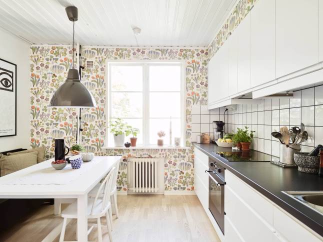 Kolorowa  kwiatowa tapeta na ścianie w białej kuchni skandynawskiej z szarą lampą i drewnianą podłogą