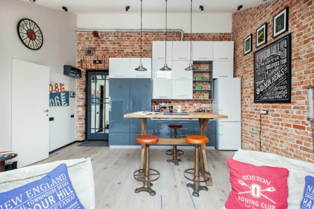 Biało-niebieska kuchnia w otwartym widoku na salon ze ścianą z czerwonej cegły