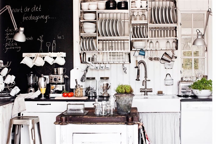 Ściana tablicowa,zestaw wiszących półek z naczyniami i biała wyspa z metalowym blatem w kuchni