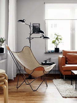Dizajnerski brązowy fotel motyl, czarna lampa podłogowa z trzema abażurami,kwadratowy czarny stolik pomocniczy z tacą,ruda skórzana sofa w salonie