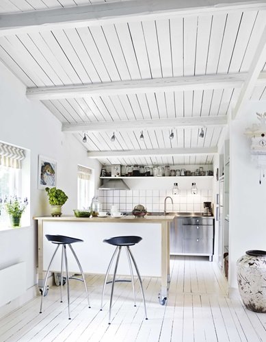 Biała kuchnia w białym domku z kuchenną wyspą na kółkach i czarnymi wysokimi stołkami barowymi na metalowych nogach