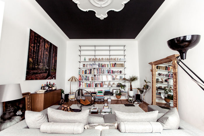 Czarny sufit z białymi ornamentem ze sztukaterii,rzeźbione lustro,nowoczesna biala sofa,i  drewniana komoda w eklektycznym salonie