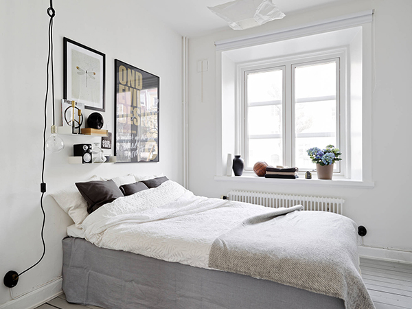 Żarówka na kablu,papierowy abażur na wiszącej lampie,nowoczesne grafiki na ścianie i biało-szara dekoracja łóżka z czarnymi poduszkami